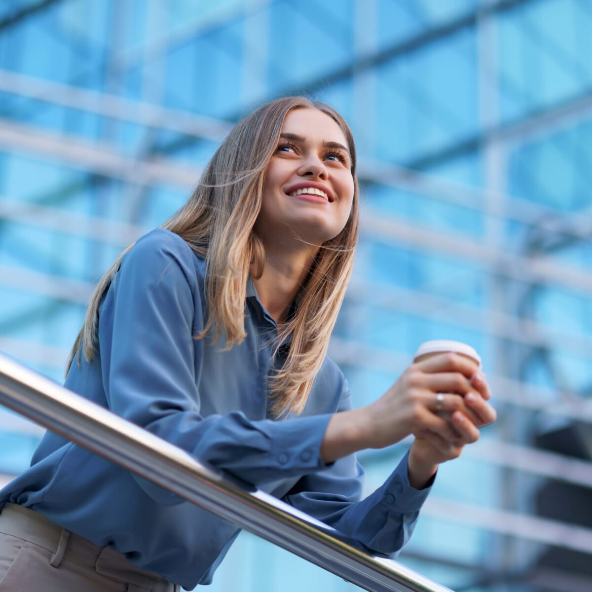Young smiling professional woman having a coffee break during her full working day. She holds a paper cup outdoors near the business building while relaxing and enjoying her beverage.